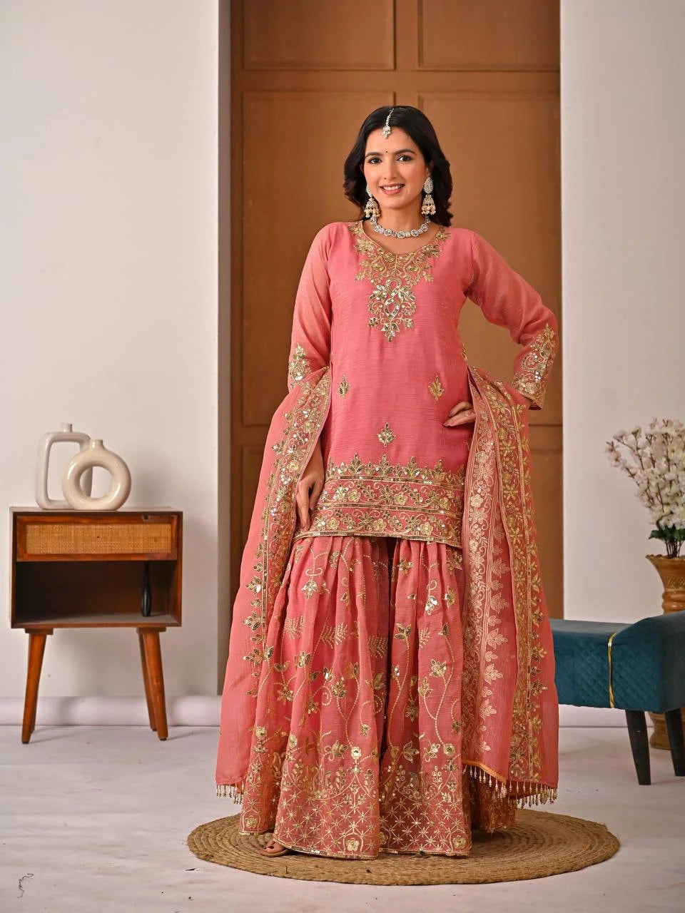 Woman in a traditional pink and gold embroidered outfit standing in a room with wooden furniture and decor.