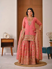 Woman in a traditional pink and gold embroidered outfit standing in a room with wooden furniture and decor.
