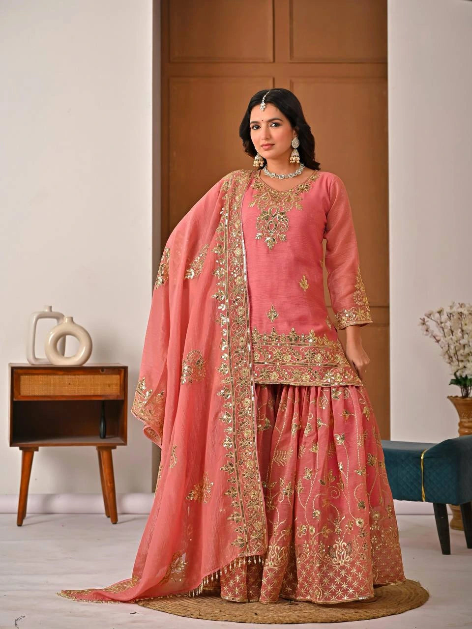 Woman in a traditional pink and gold embroidered outfit standing in a room with wooden furniture and decor.