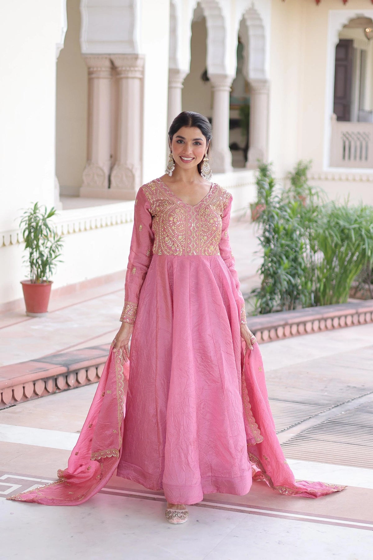 Woman in a pink traditional outfit standing in front of a building with columns and plants.