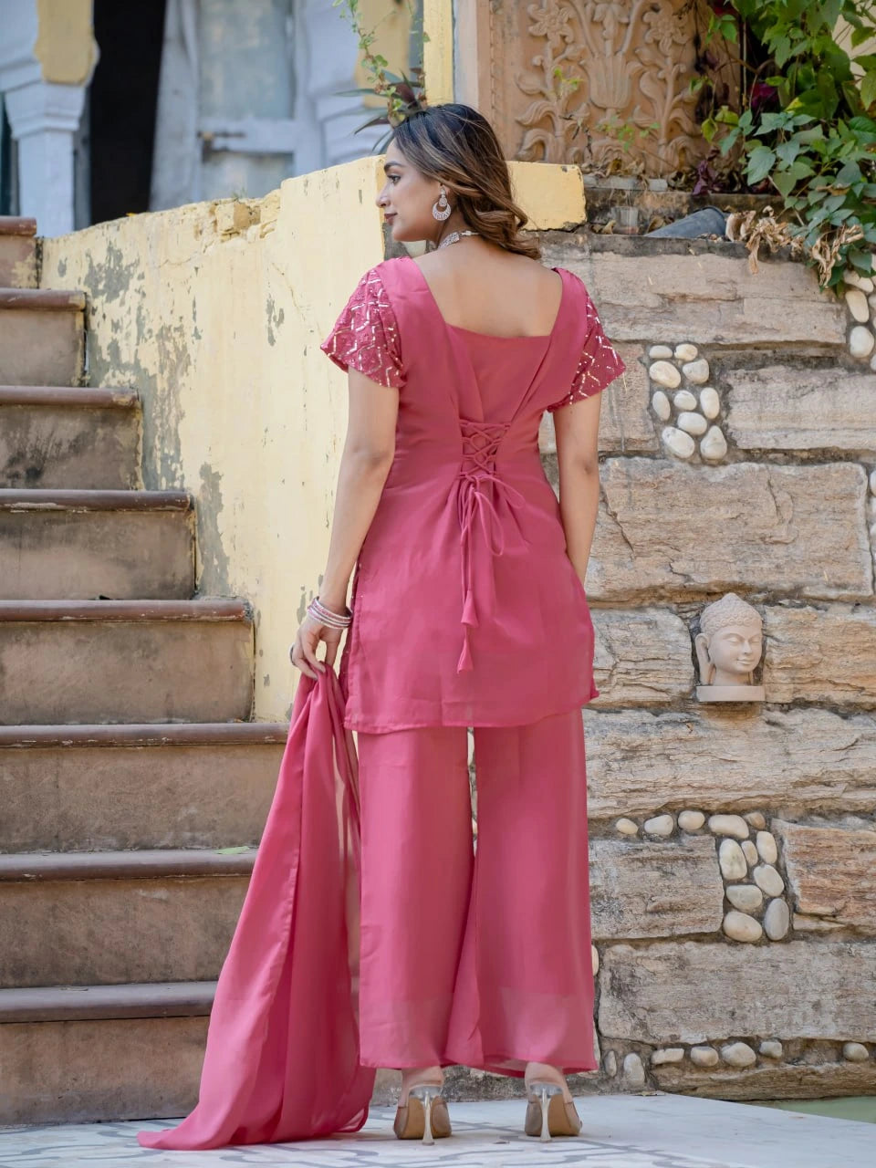 Woman in a pink outfit standing on stone steps with a stone wall and plants in the background