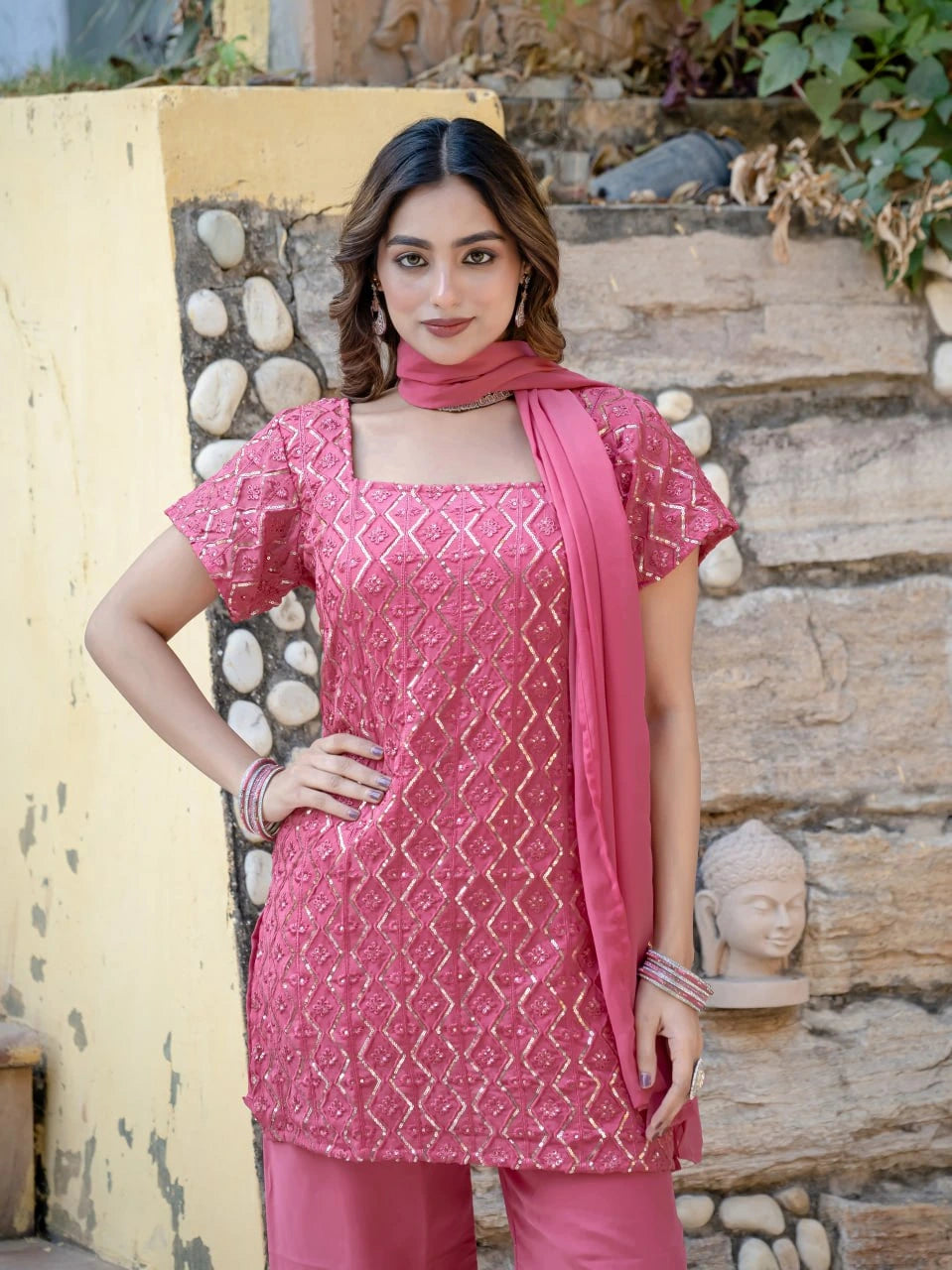 Woman wearing a pink traditional outfit with a patterned top and matching dupatta, standing against a stone wall.