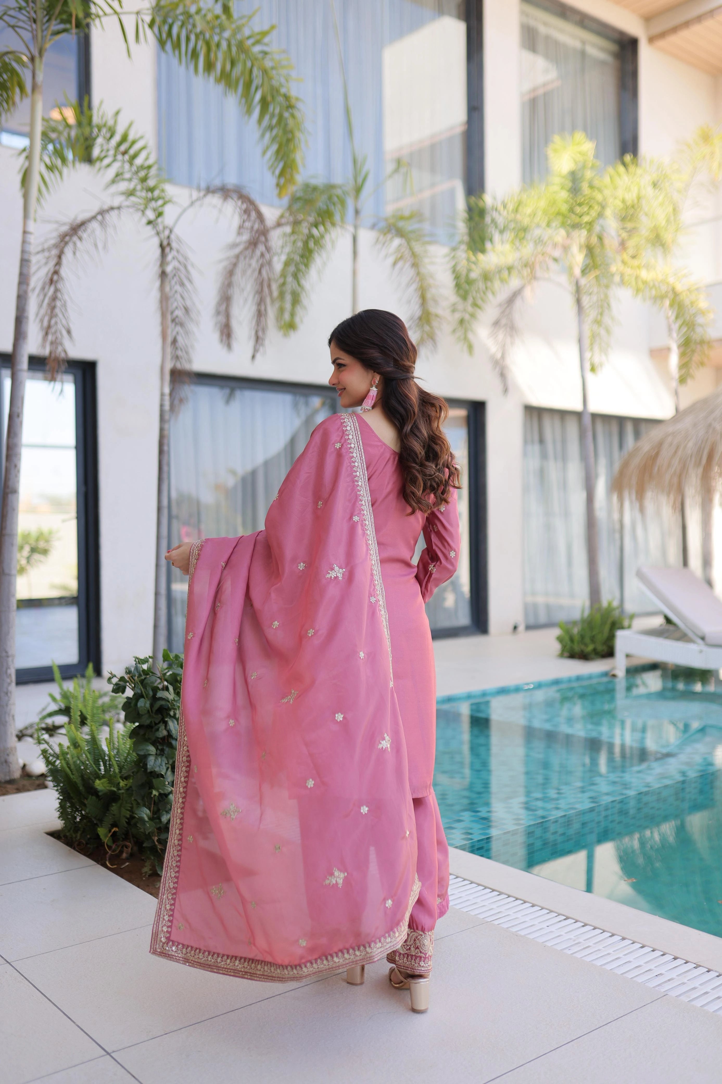 Woman in a pink traditional outfit standing by a pool with modern building and plants in the background