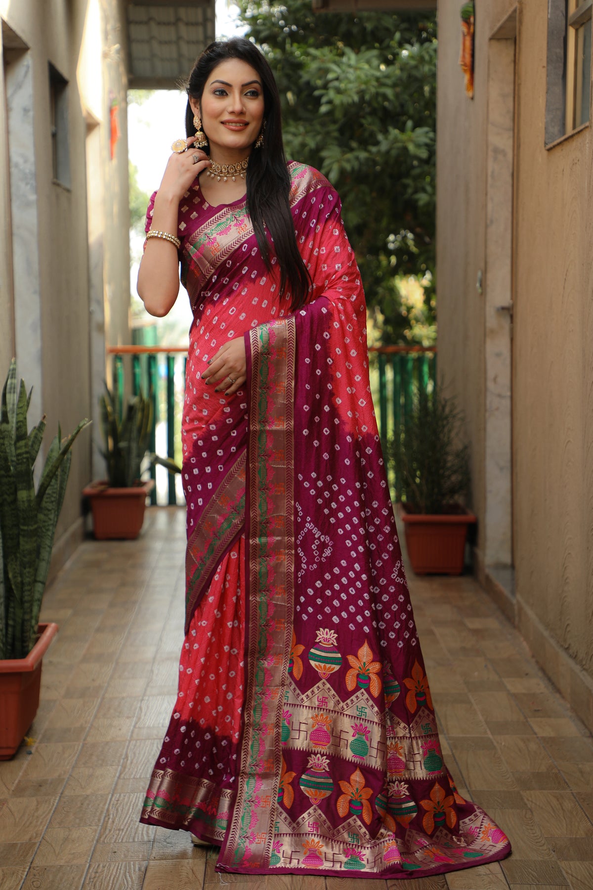 Woman in a red and purple saree standing in an outdoor setting with plants.