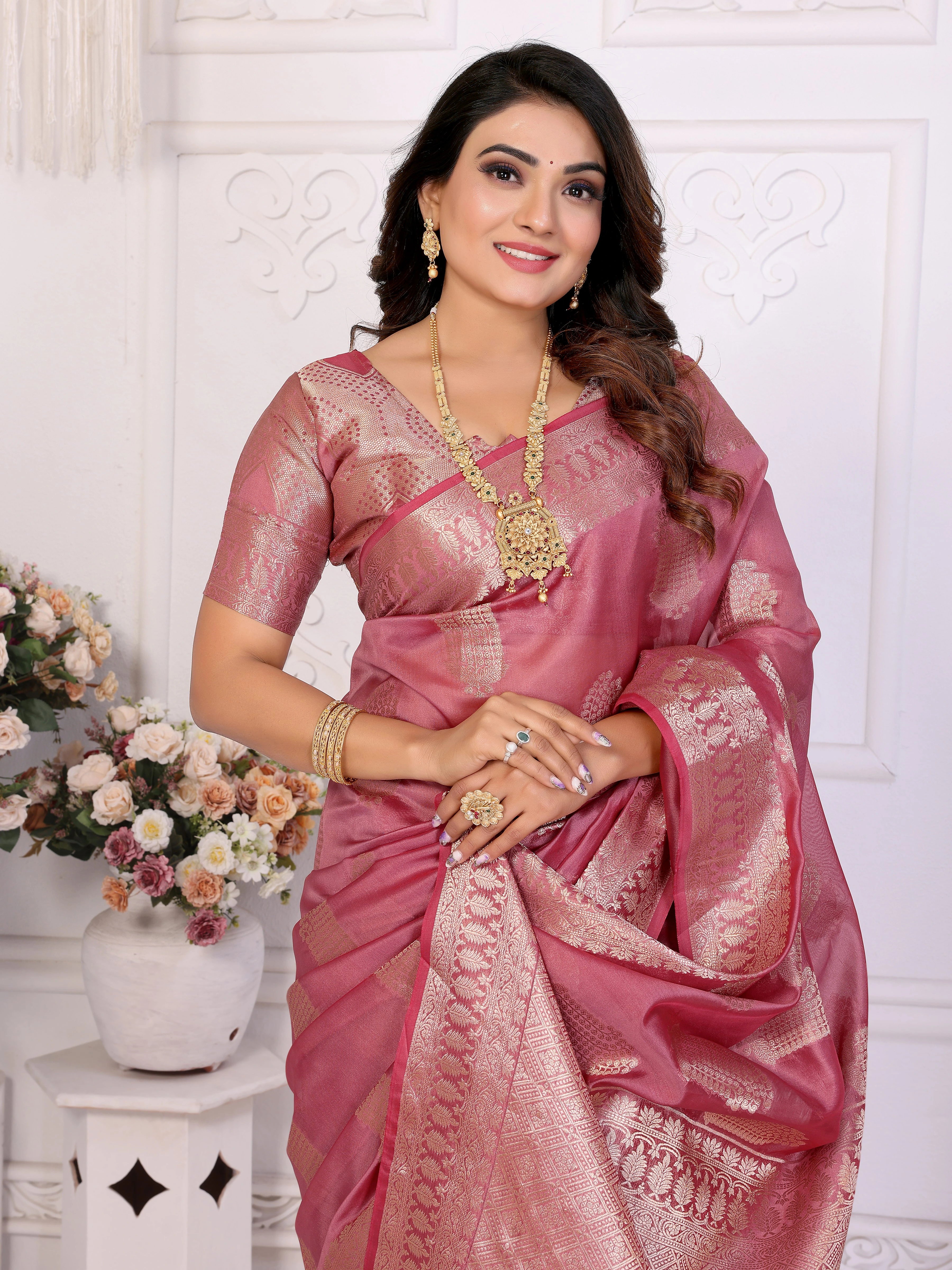 Woman in a pink saree with gold jewelry against a white wall