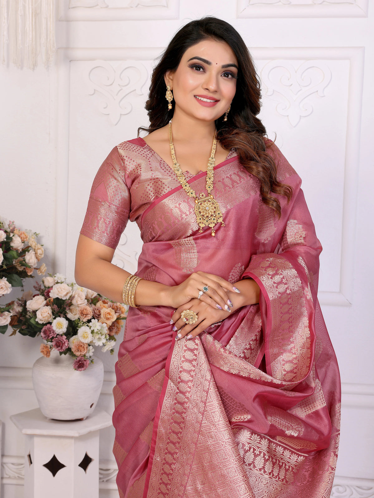 Woman in a pink saree with gold jewelry against a white wall