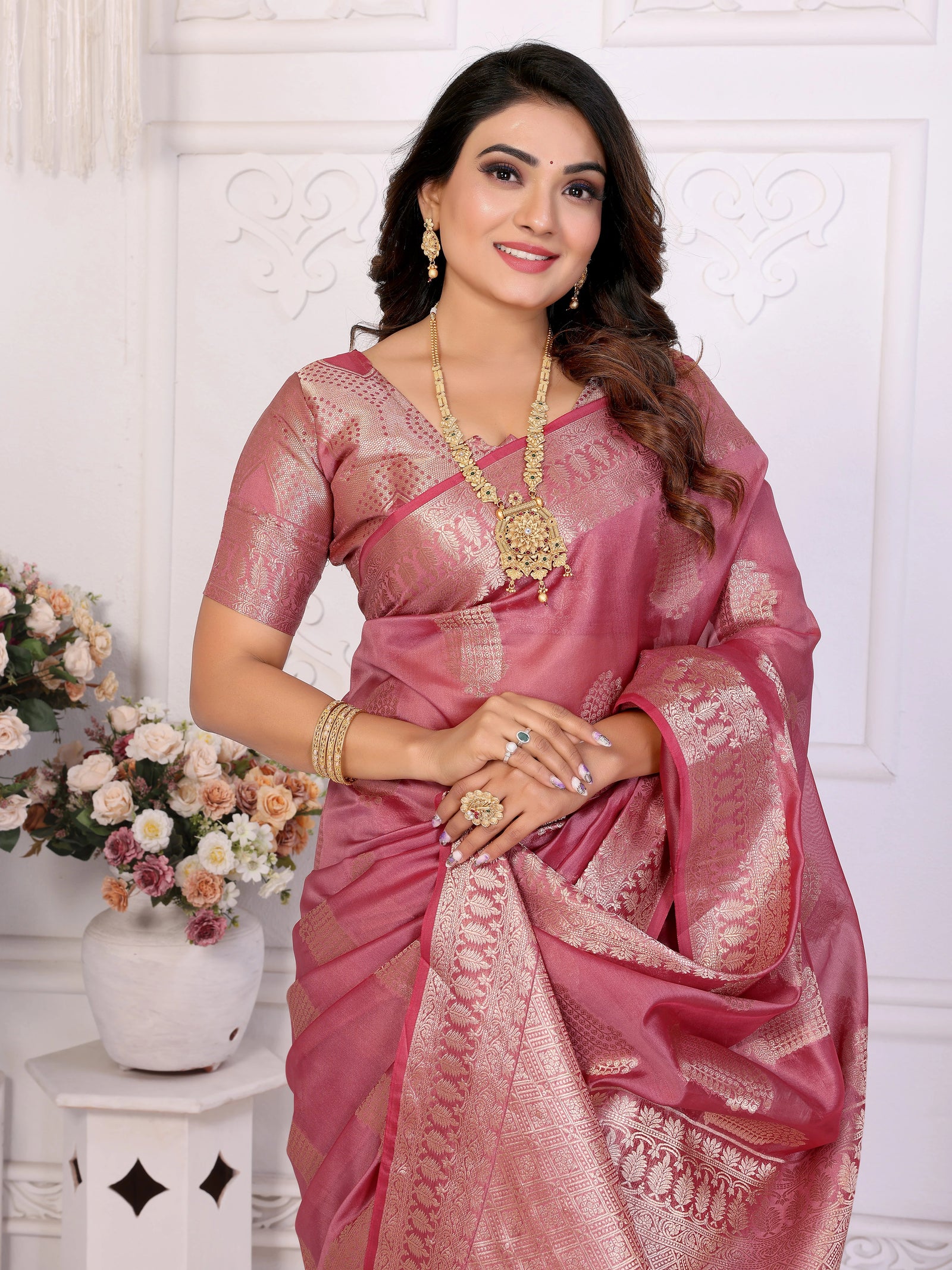 Woman in a pink saree with gold jewelry against a white wall