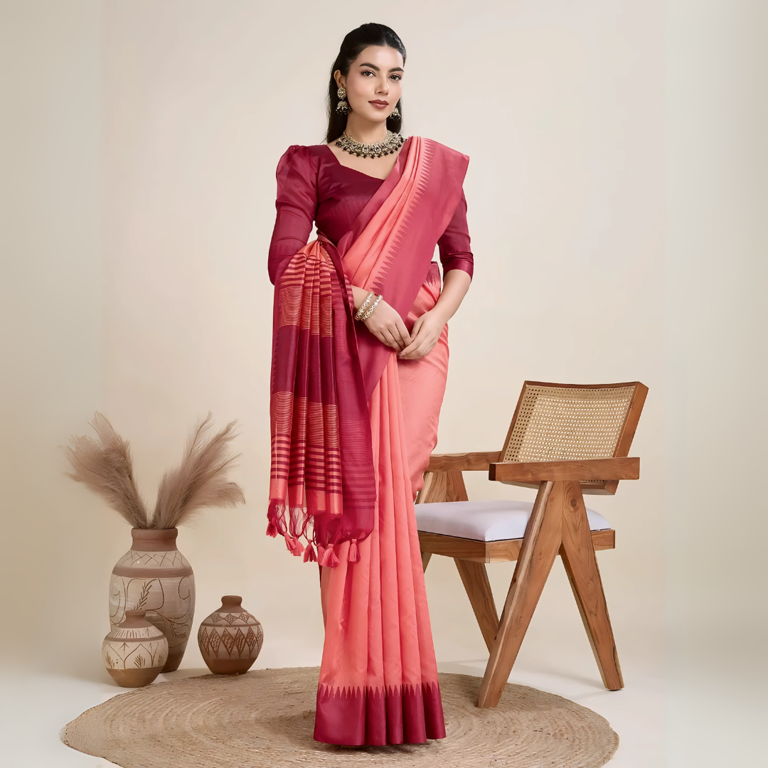 A woman models a striking two-toned Raw Silk Saree. The main body is Coral Pink, and the pallu/border contrast dramatically in Deep Maroon/Wine Red. The saree features traditional temple weaving and horizontal stripe patterns on the pallu. She wears a long-sleeve maroon blouse and accessorizes with a chunky dark gemstone necklace, posing indoors.