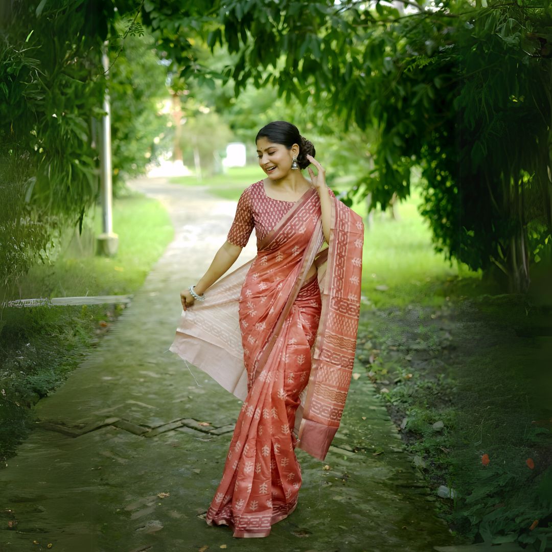 A woman models a Terracotta Red Soft Cotton Saree featuring an all-over digital block print of traditional motifs. She wears a printed red blouse and silver jhumka earrings, posing on an outdoor path framed by greenery.