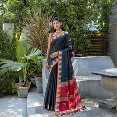 A woman models a dramatic Black Tussar Silk Saree with small red woven motifs. The saree has a beige temple-style border and a pallu featuring wide bands of red and beige weaving. She wears a red sleeveless blouse and a gold choker necklace.