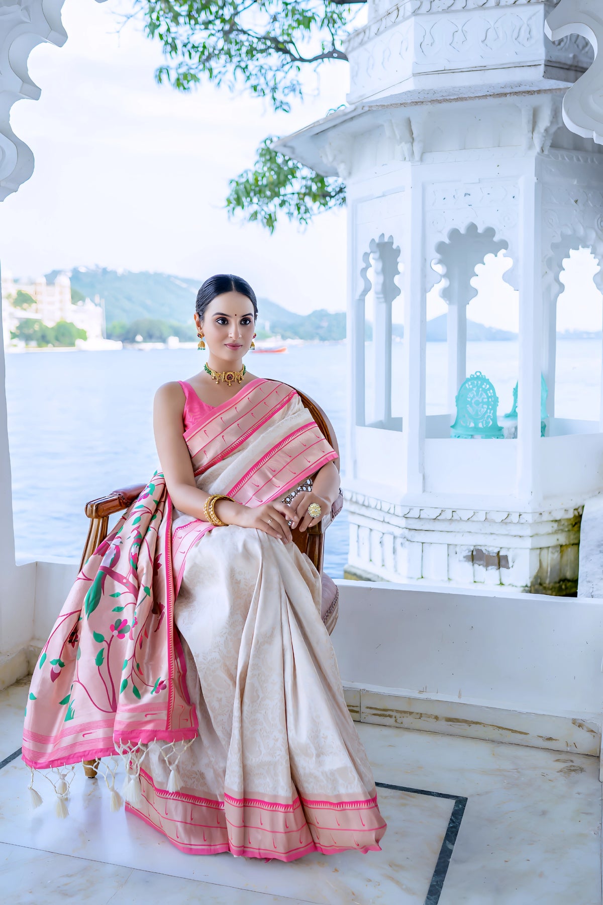 A woman models an elegant White or Cream Soft Silk Saree. The saree features a subtle tone-on-tone weave and a striking pink contrast border with thin gold Zari. The pallu is a focal point, featuring rich, artistic Paithani-style print or weaving of pink, red, and green bird-on-branch motifs. She wears a vibrant pink sleeveless blouse and gold Kundan jewelry, posing outdoors by a lake.