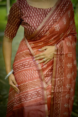 A woman models a Terracotta Red Soft Cotton Saree featuring an all-over digital block print of traditional motifs. She wears a printed red blouse and silver jhumka earrings, posing on an outdoor path framed by greenery.