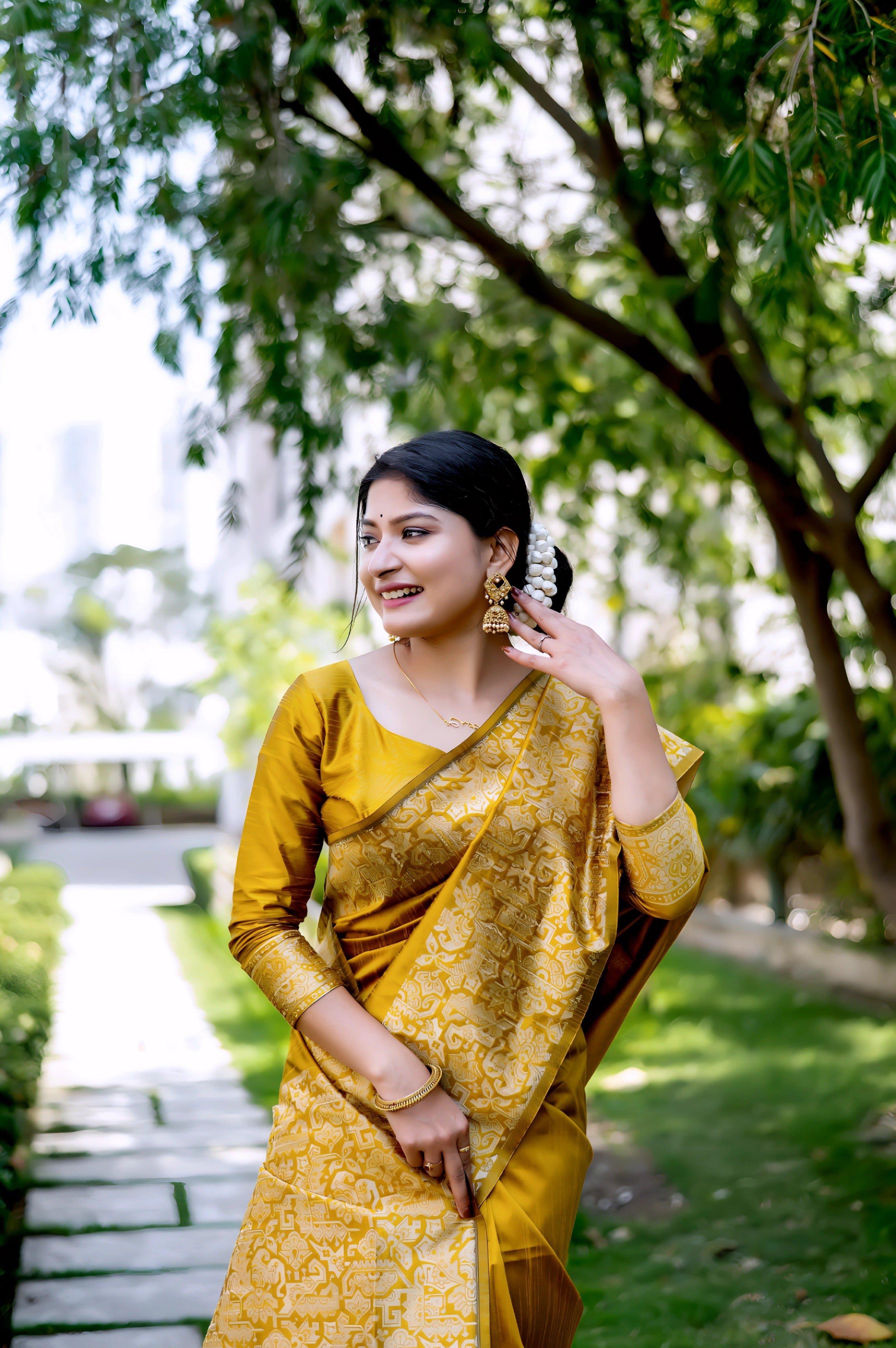 A woman models a luxurious Golden Yellow Raw Silk Saree. The saree features a wide border and pallu with dense, tone-on-tone gold Zari weaving in a Kalamkari-style floral and paisley pattern. She wears a matching long-sleeve yellow blouse and traditional gold jewelry, including a maang tikka, posing outdoors on a sunlit garden path.