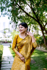 A woman models a luxurious Golden Yellow Raw Silk Saree. The saree features a wide border and pallu with dense, tone-on-tone gold Zari weaving in a Kalamkari-style floral and paisley pattern. She wears a matching long-sleeve yellow blouse and traditional gold jewelry, including a maang tikka, posing outdoors on a sunlit garden path.