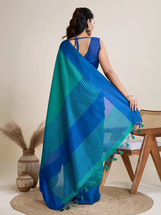 A woman models a vibrant Two-Toned Sea Blue and Royal Blue Raw Silk Saree. The saree is cinched at the waist with a black leather belt with a circular buckle. She wears a blue sleeveless blouse and accessorizes with chunky tribal jewelry, posing indoors.