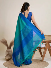 A woman models a vibrant Two-Toned Sea Blue and Royal Blue Raw Silk Saree. The saree is cinched at the waist with a black leather belt with a circular buckle. She wears a blue sleeveless blouse and accessorizes with chunky tribal jewelry, posing indoors.
