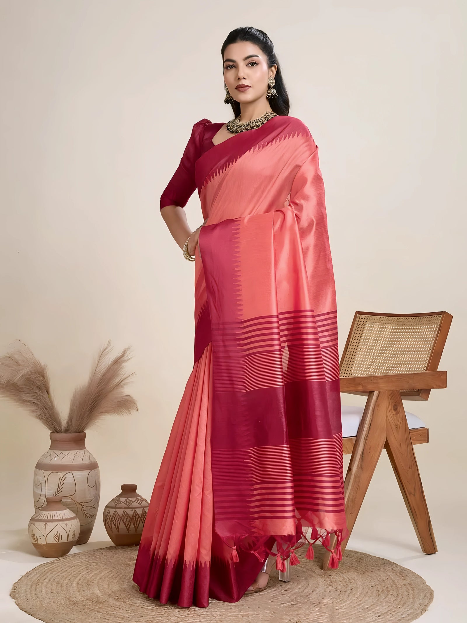 A woman models a striking two-toned Raw Silk Saree. The main body is Coral Pink, and the pallu/border contrast dramatically in Deep Maroon/Wine Red. The saree features traditional temple weaving and horizontal stripe patterns on the pallu. She wears a long-sleeve maroon blouse and accessorizes with a chunky dark gemstone necklace, posing indoors.