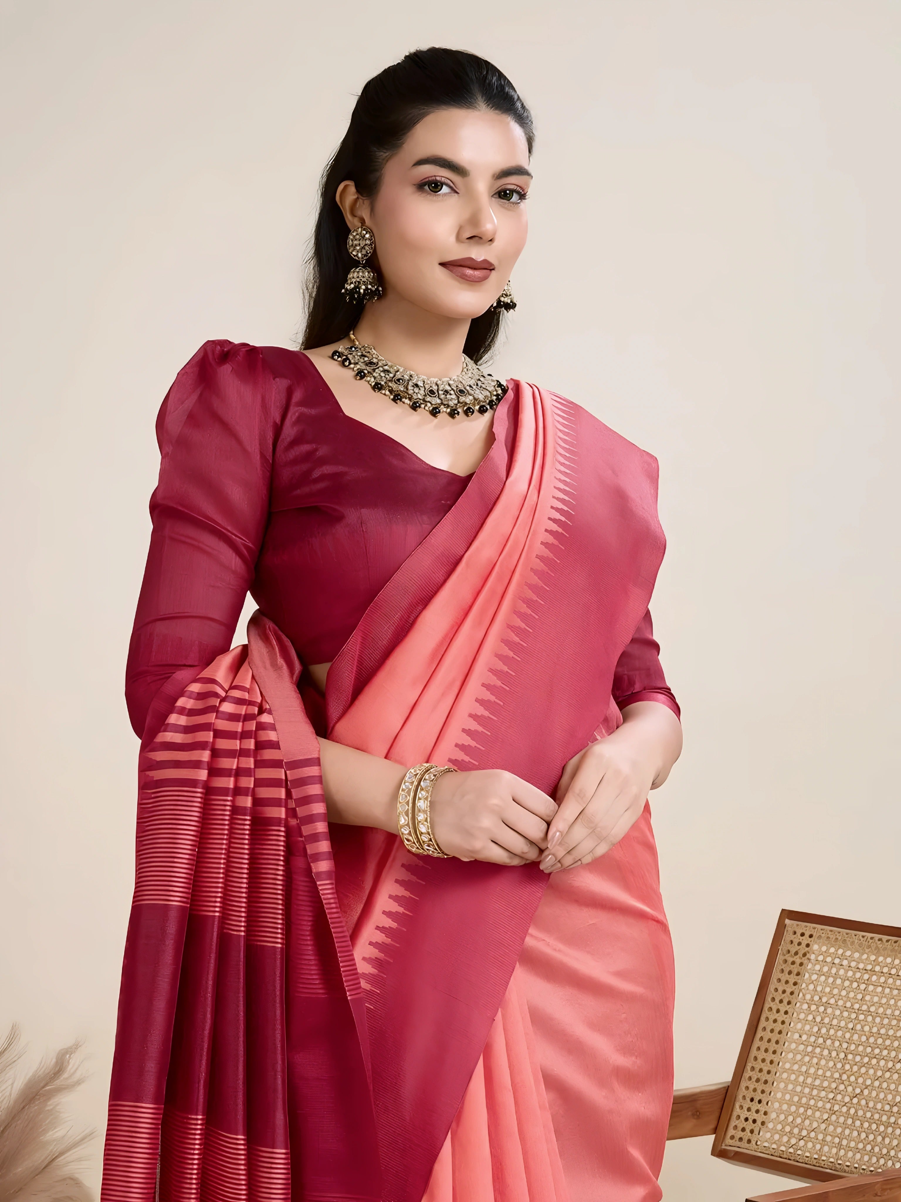A woman models a striking two-toned Raw Silk Saree. The main body is Coral Pink, and the pallu/border contrast dramatically in Deep Maroon/Wine Red. The saree features traditional temple weaving and horizontal stripe patterns on the pallu. She wears a long-sleeve maroon blouse and accessorizes with a chunky dark gemstone necklace, posing indoors.