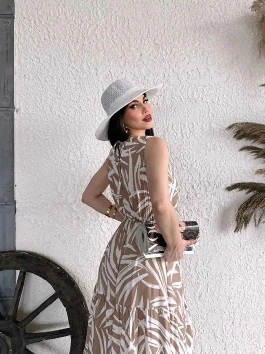 Woman in a patterned dress and white hat standing against a textured white wall.