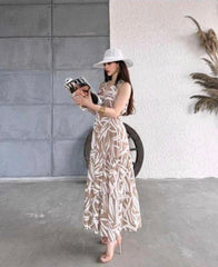 Woman in a long patterned dress and hat holding a book against a gray wall.