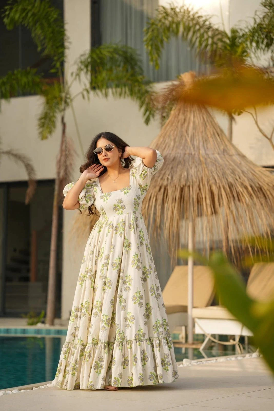 Woman in a floral dress standing by a poolside with a thatched umbrella in the background
