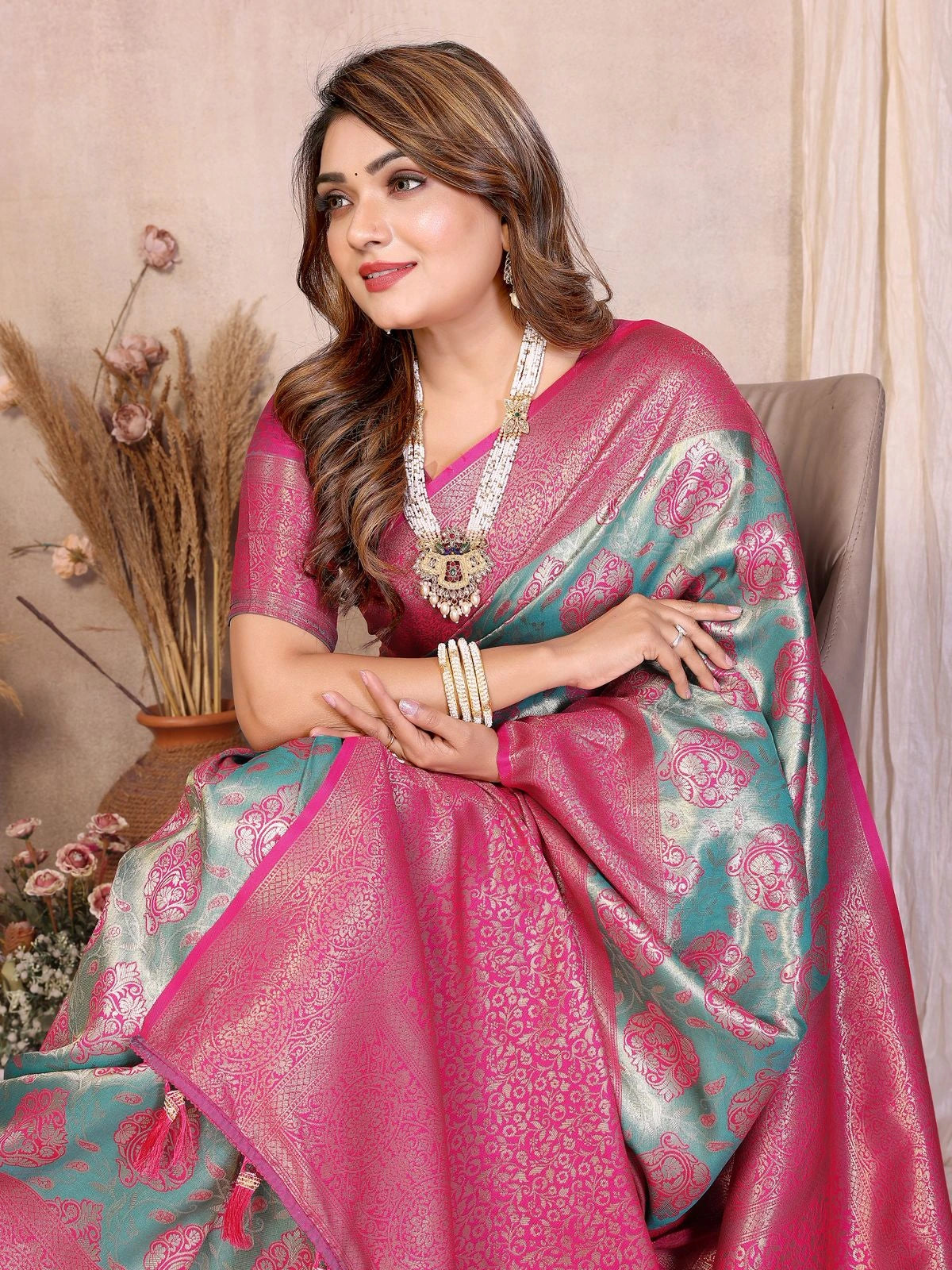 Woman wearing a pink and green saree with jewelry, sitting indoors.