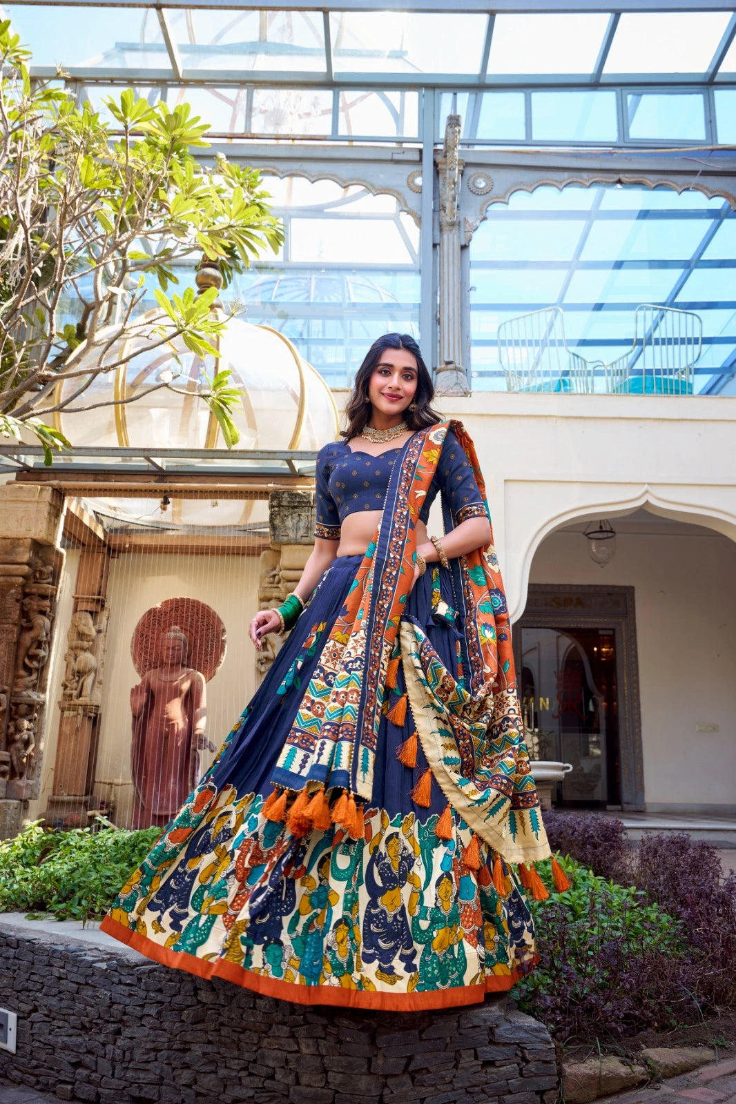 Woman in a colorful traditional outfit standing in an outdoor setting with plants and architectural elements.