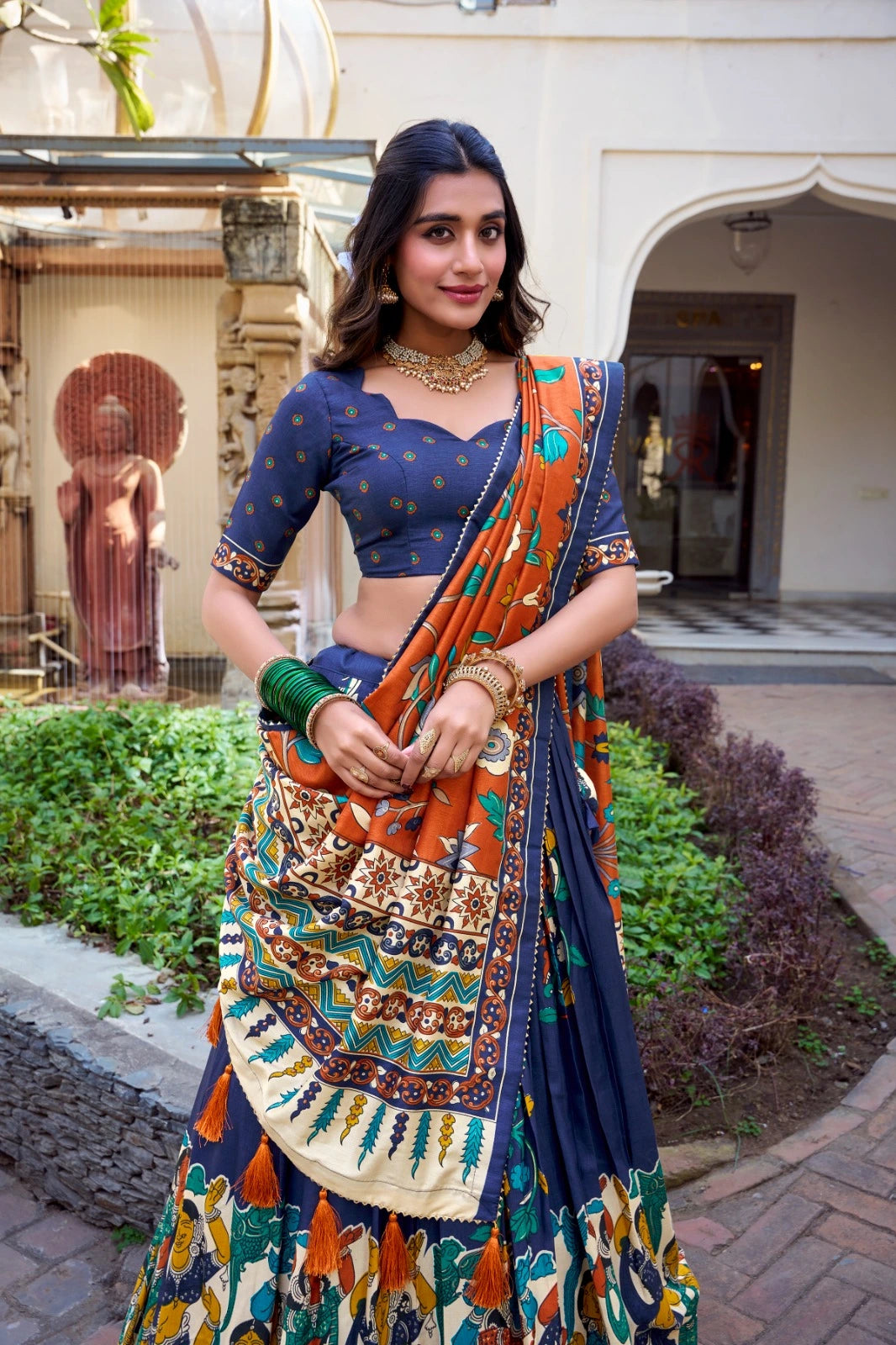 Woman in a colorful saree standing outdoors with a building and statue in the background