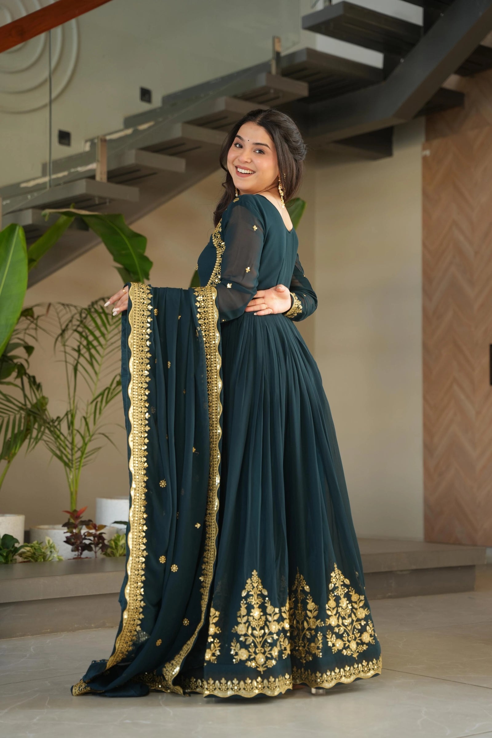 Woman in a teal saree with gold embroidery standing indoors.
