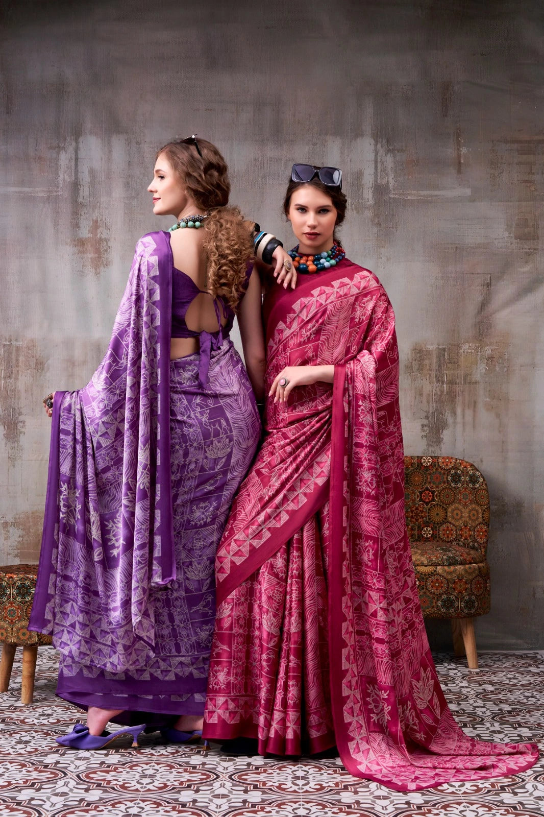 Two women in vibrant purple and pink sarees standing against a textured wall.