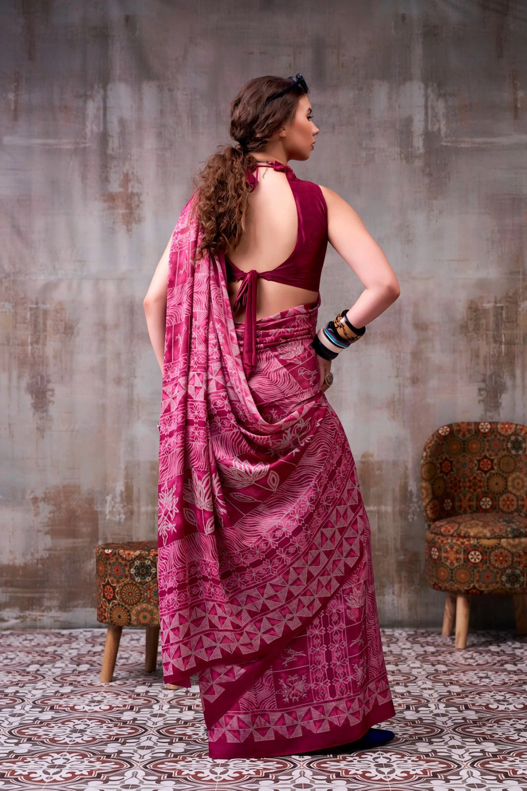 Woman in a pink saree with a maroon blouse standing against a textured wall.