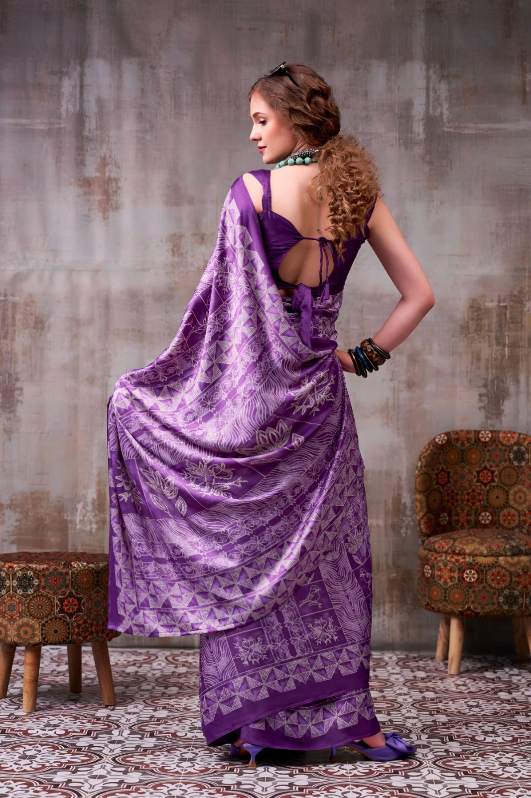 Woman in a purple saree with a textured wall and patterned chair in the background