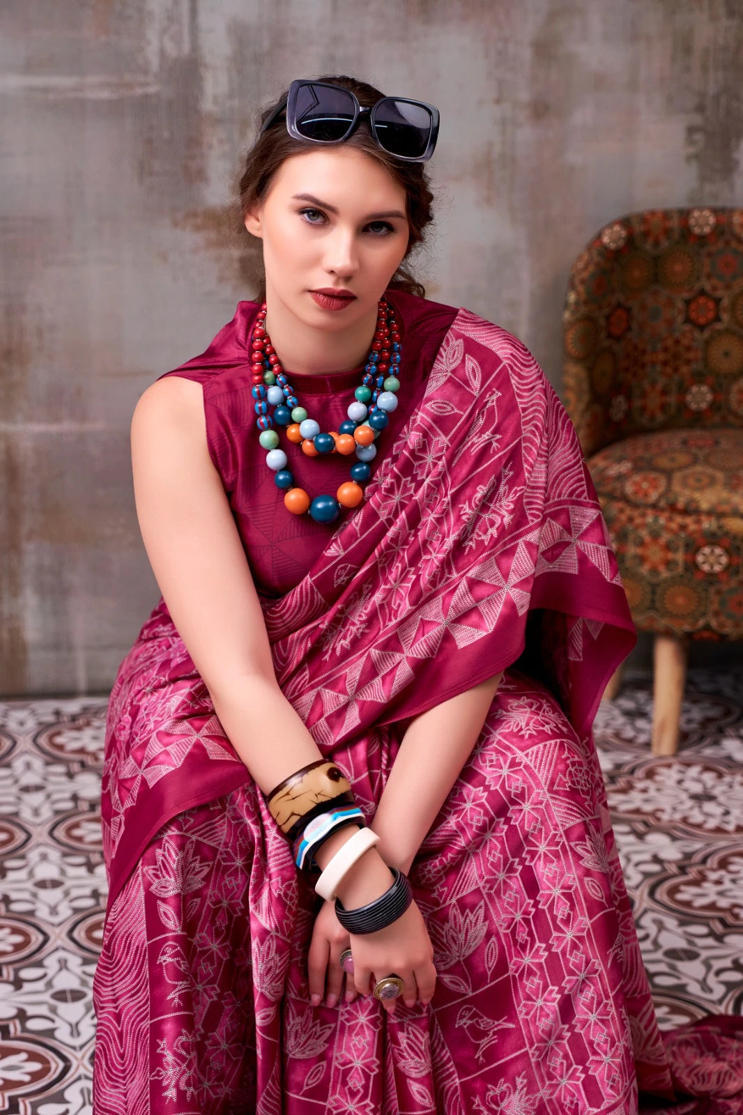 Woman in a pink saree with colorful jewelry sitting on a patterned rug.