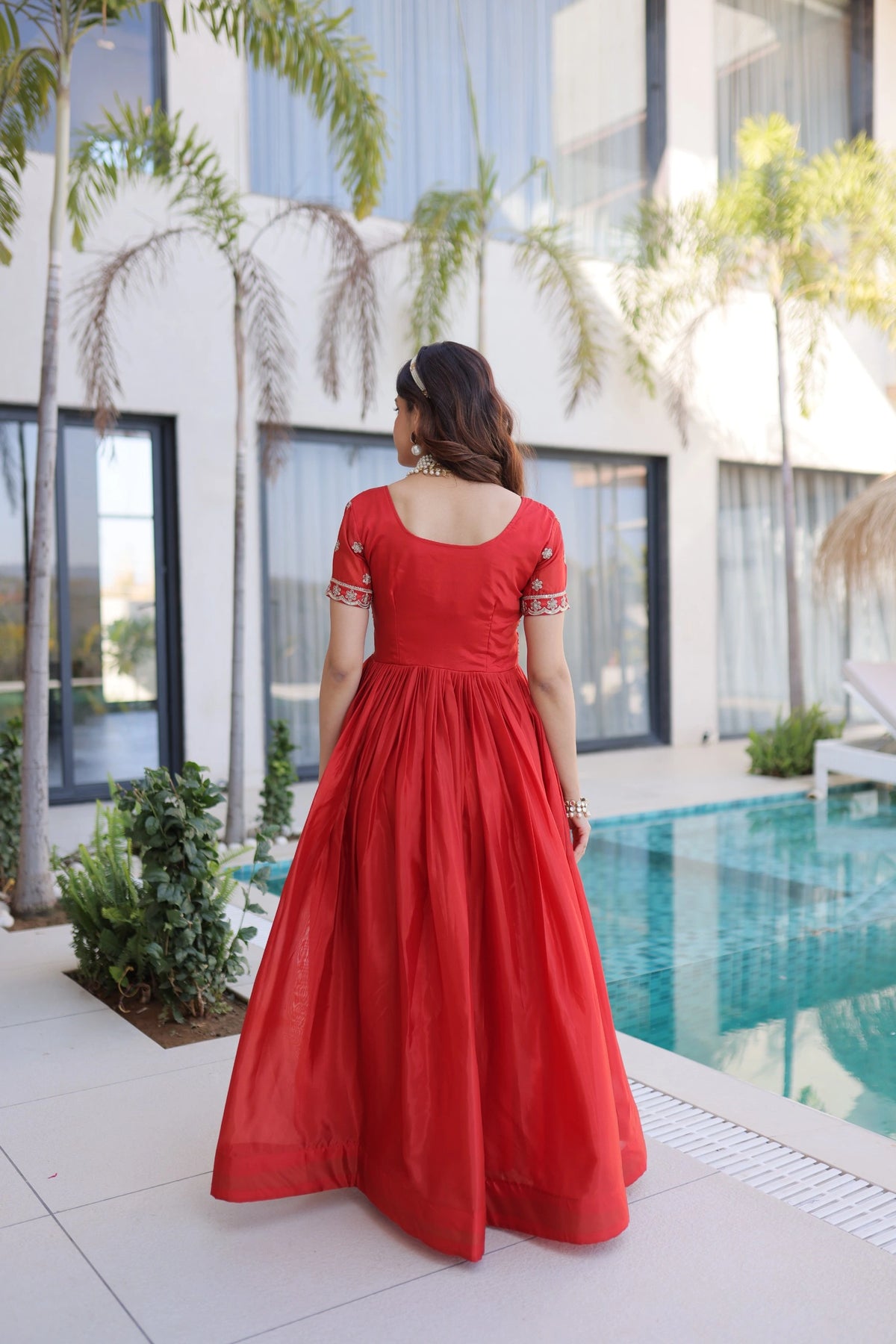 Woman in a red dress standing by a pool with a modern building and palm trees in the background