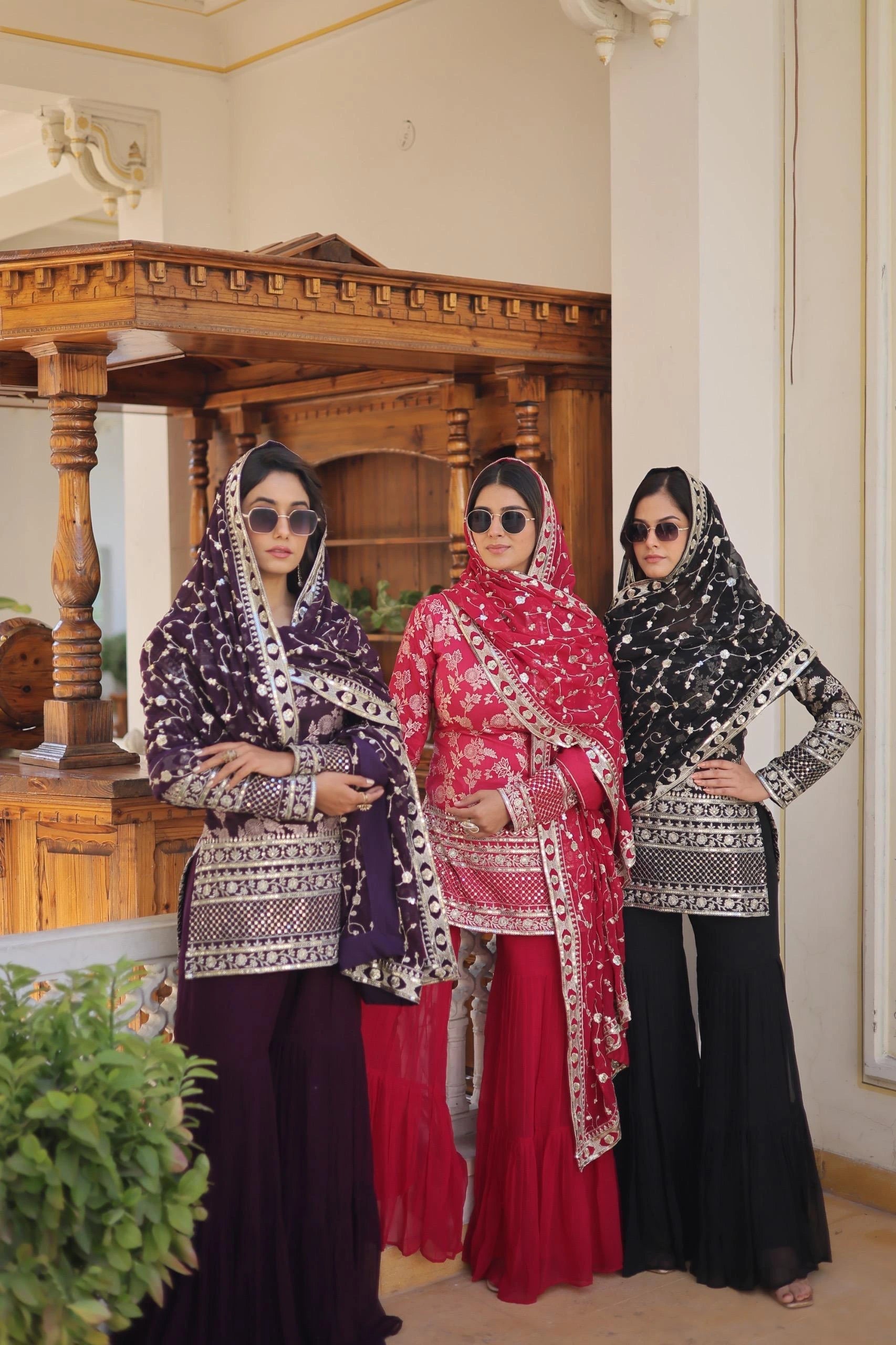 Three women in traditional attire standing indoors with a wooden structure and decorative elements in the background.