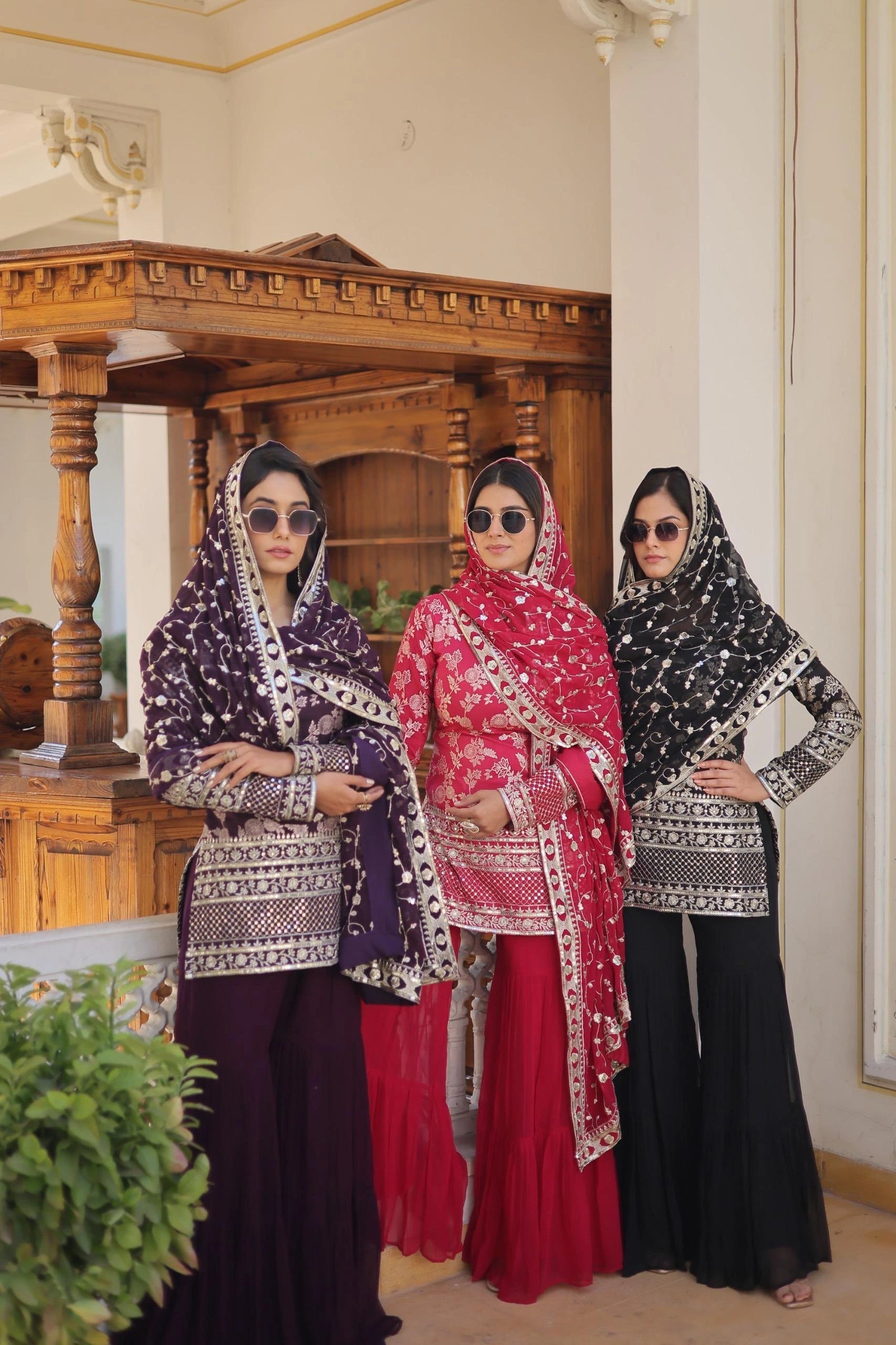 Three women in traditional attire standing indoors with a wooden structure and decorative elements in the background.