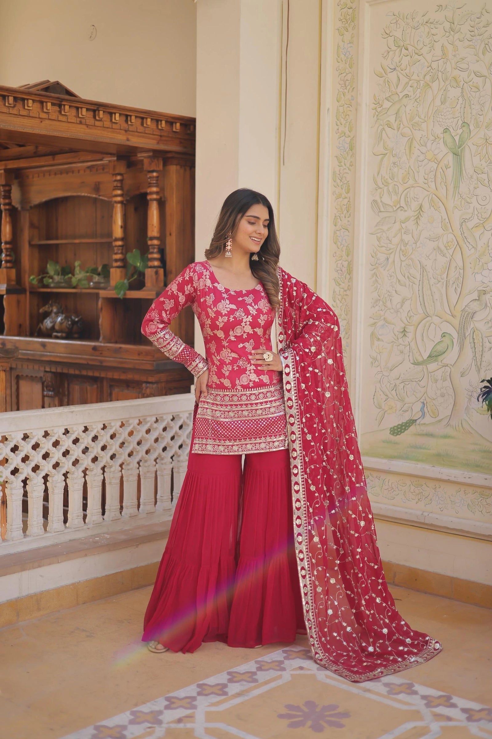 Woman in a traditional pink and red outfit standing in an ornate room.