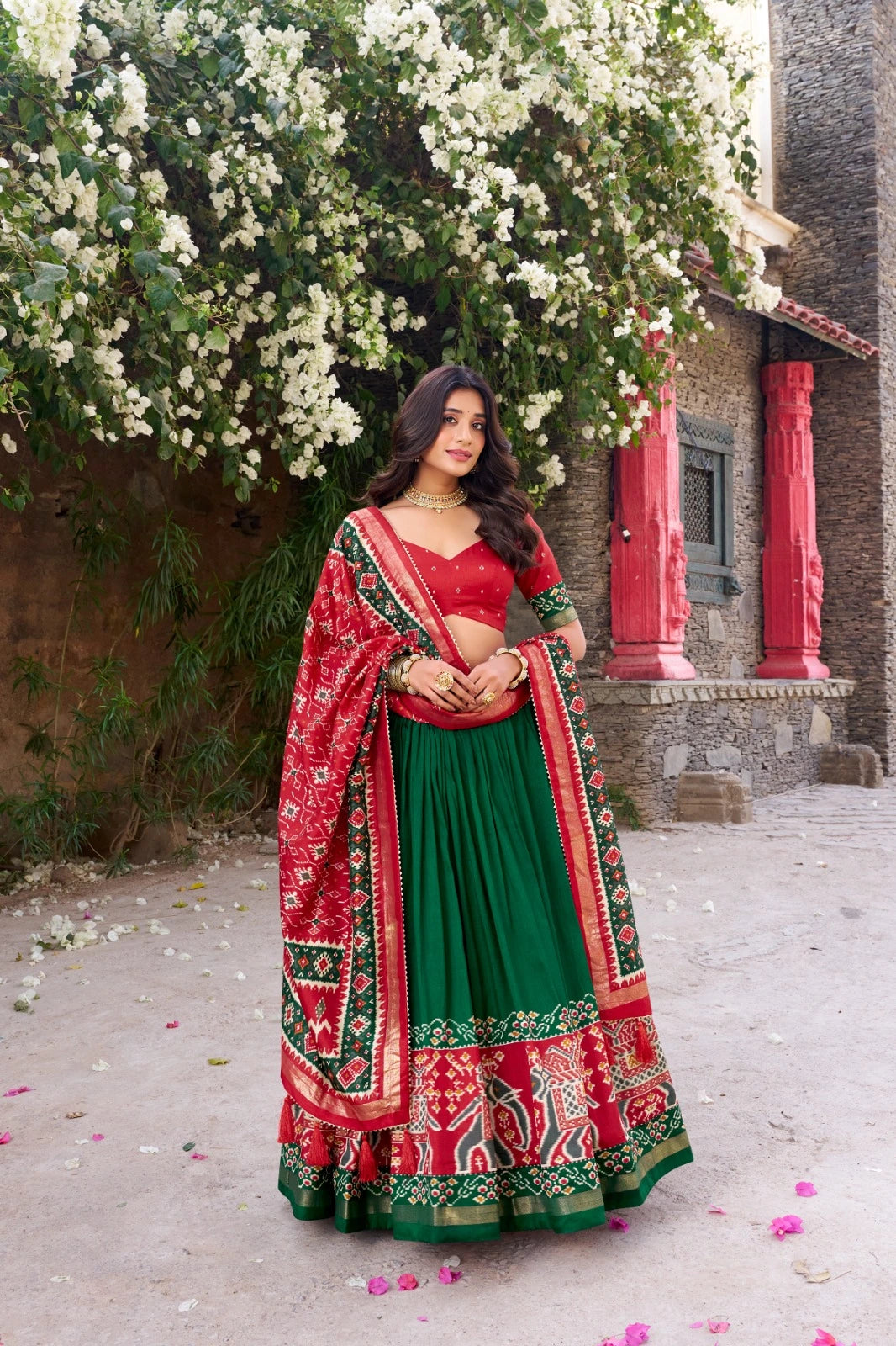 Woman in a red and green traditional outfit standing in front of a building with white flowers.