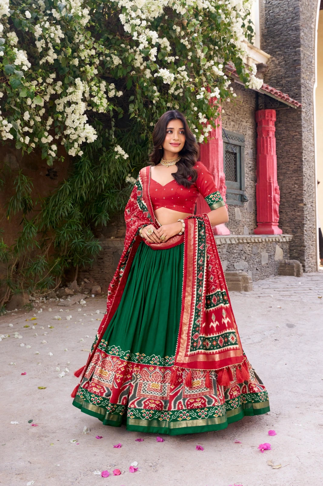 Woman in a red and green traditional outfit standing outdoors with flowers and a building in the background