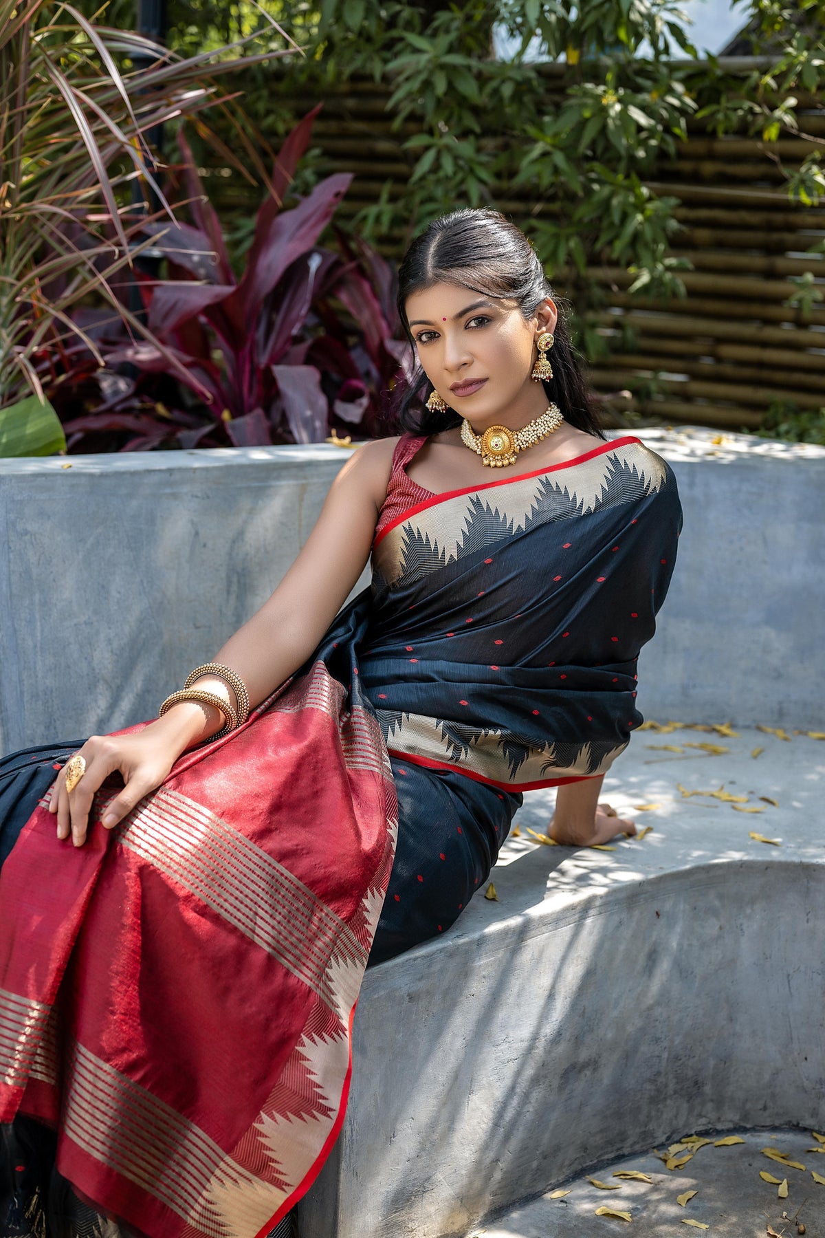 A woman models a dramatic Black Tussar Silk Saree with small red woven motifs. The saree has a beige temple-style border and a pallu featuring wide bands of red and beige weaving. She wears a red sleeveless blouse and a gold choker necklace.