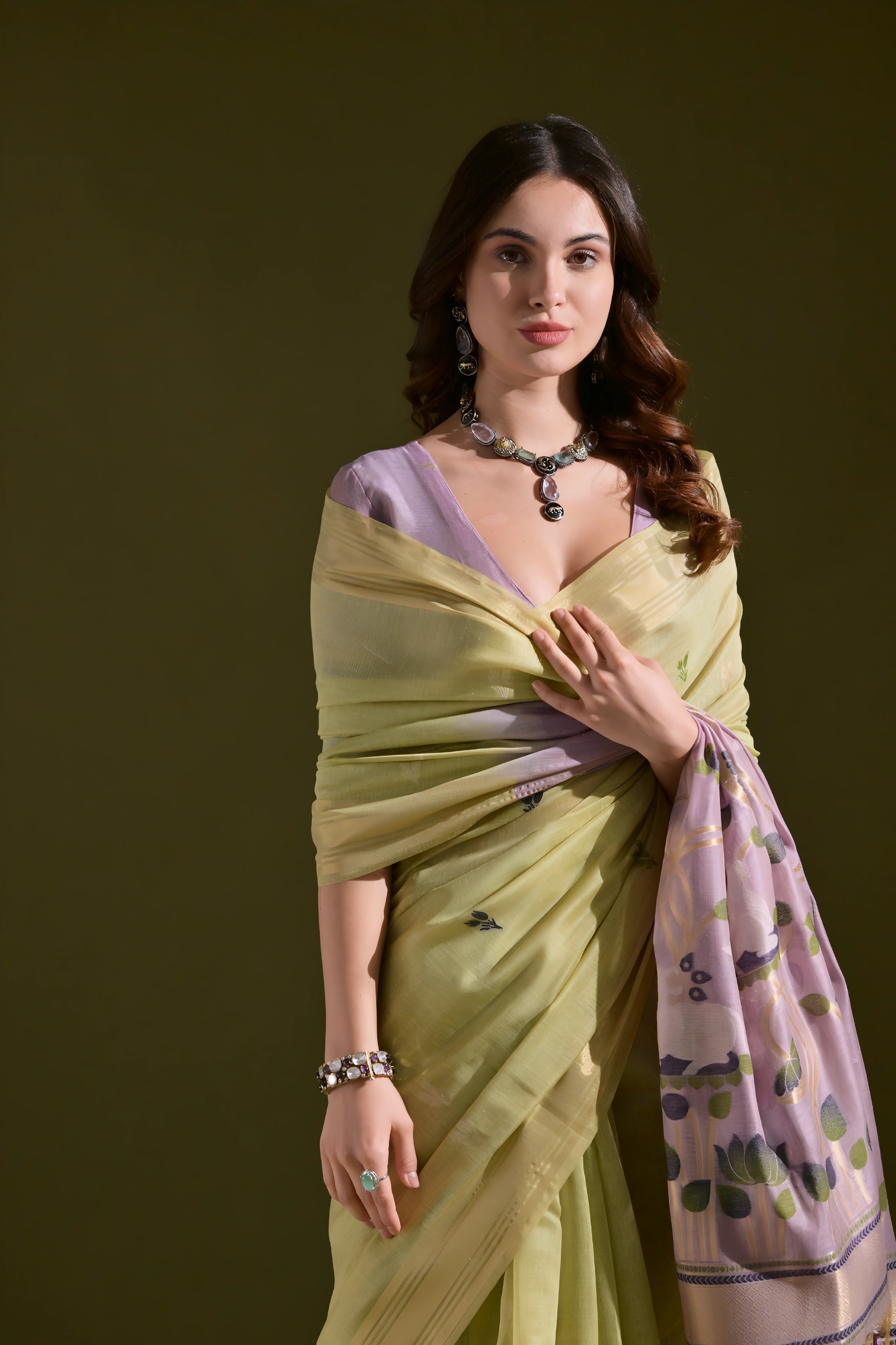A woman models a Soft Muga Cotton Saree. The saree's body is soft light green with a gold Zari border. The pallu transitions into a light purple color featuring large, artistic woven motifs in green and purple. She wears a lavender blouse and a dark choker necklace.