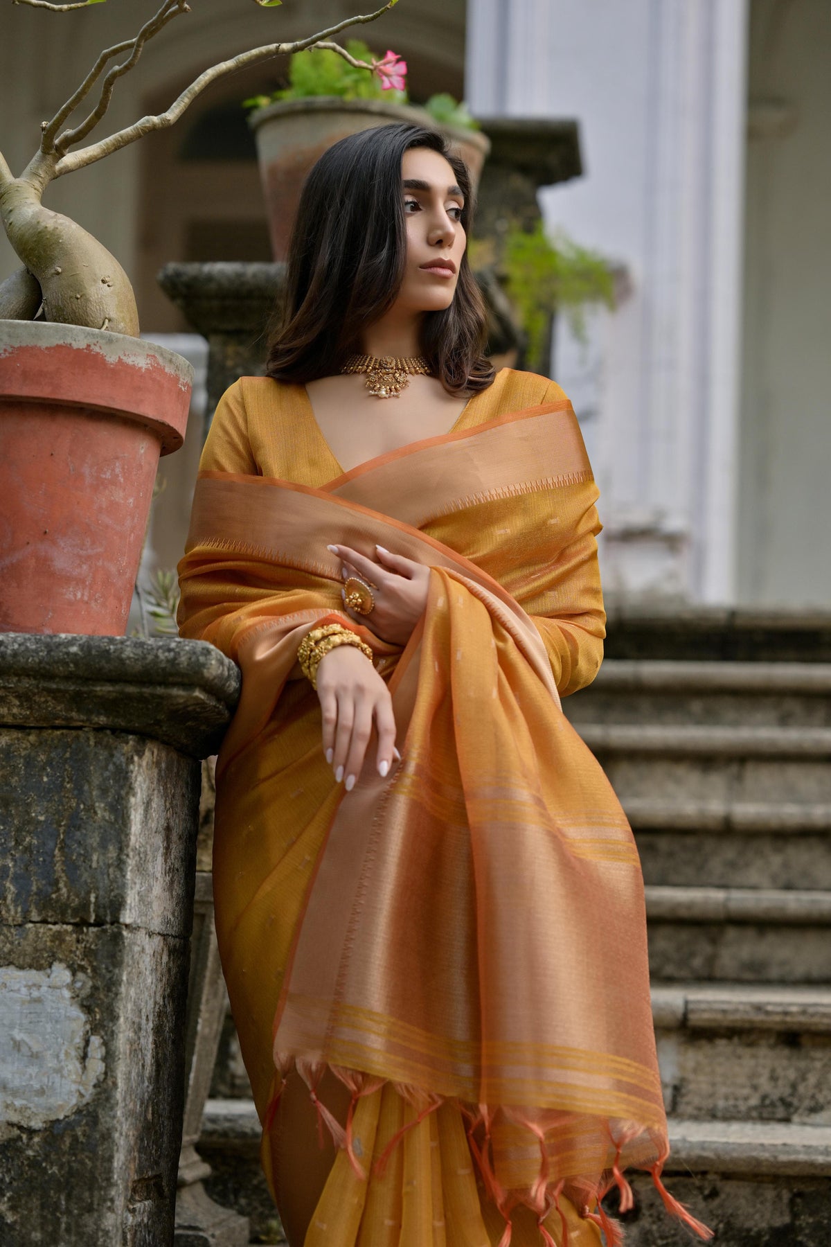 A woman models a stunning Golden Yellow Soft Silk Saree. The saree features subtle metallic motifs on the body and a wide, contrasting Copper/Rust Zari woven border. The lightweight fabric flows elegantly as she poses on a weathered stone staircase, wearing a delicate gold choker necklace.