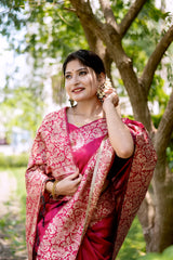 A woman models a luxurious Deep Pink Raw Silk Saree. The saree features a wide border and pallu with dense, intricate gold Zari weaving in a Kalamkari-style floral and paisley pattern. She wears a matching short-sleeve pink blouse and traditional gold jewelry, posing outdoors on a stone pathway, framed by lush greenery.
