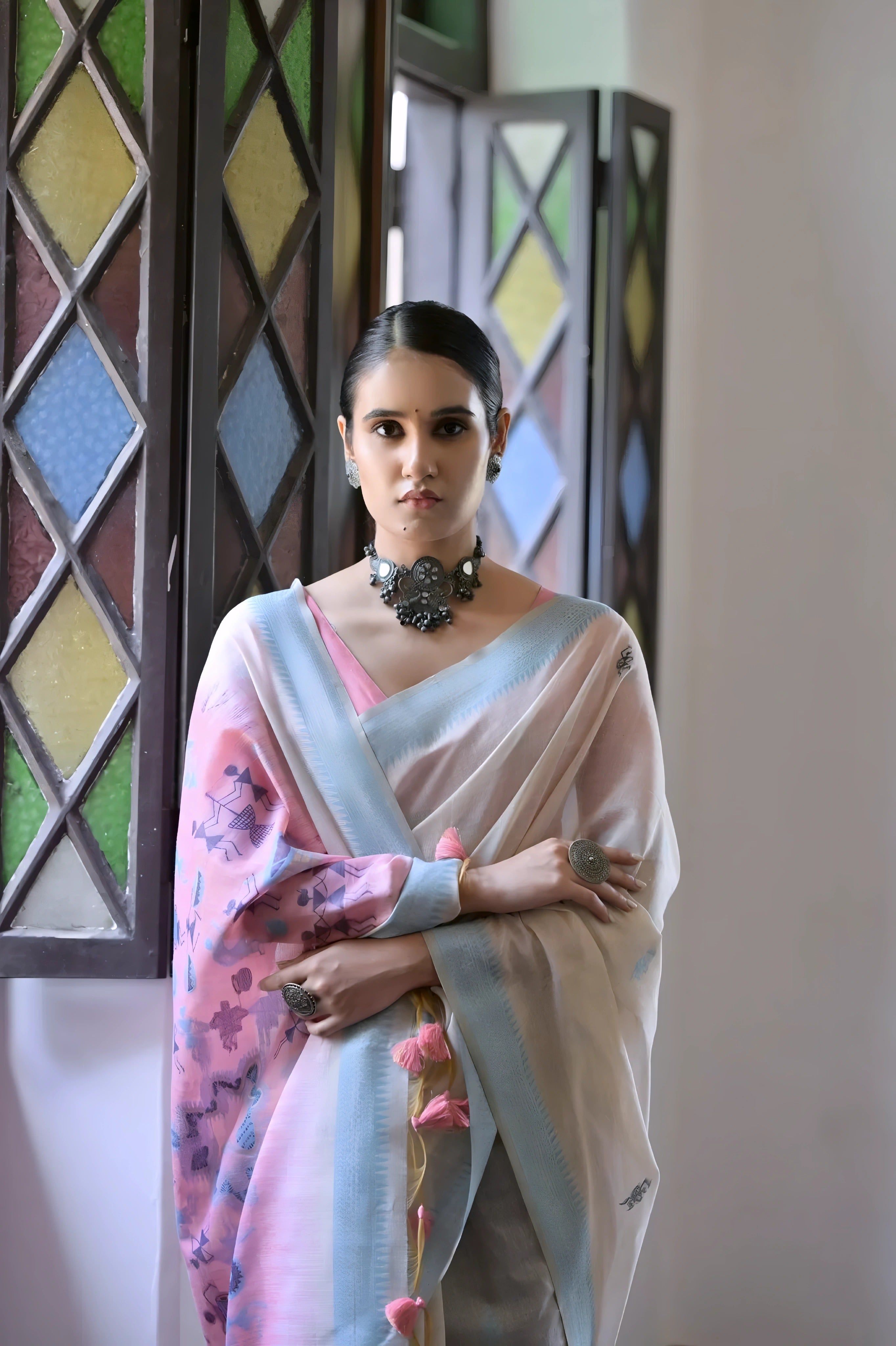 A woman models a graceful Off White Soft Muga Cotton Silk Saree. The saree has a light blue border, and the pallu features a wide pink panel decorated with traditional Warli-style figurative motifs in blue and purple. She wears a pink blouse and a dark oxidized choker necklace, posing indoors.