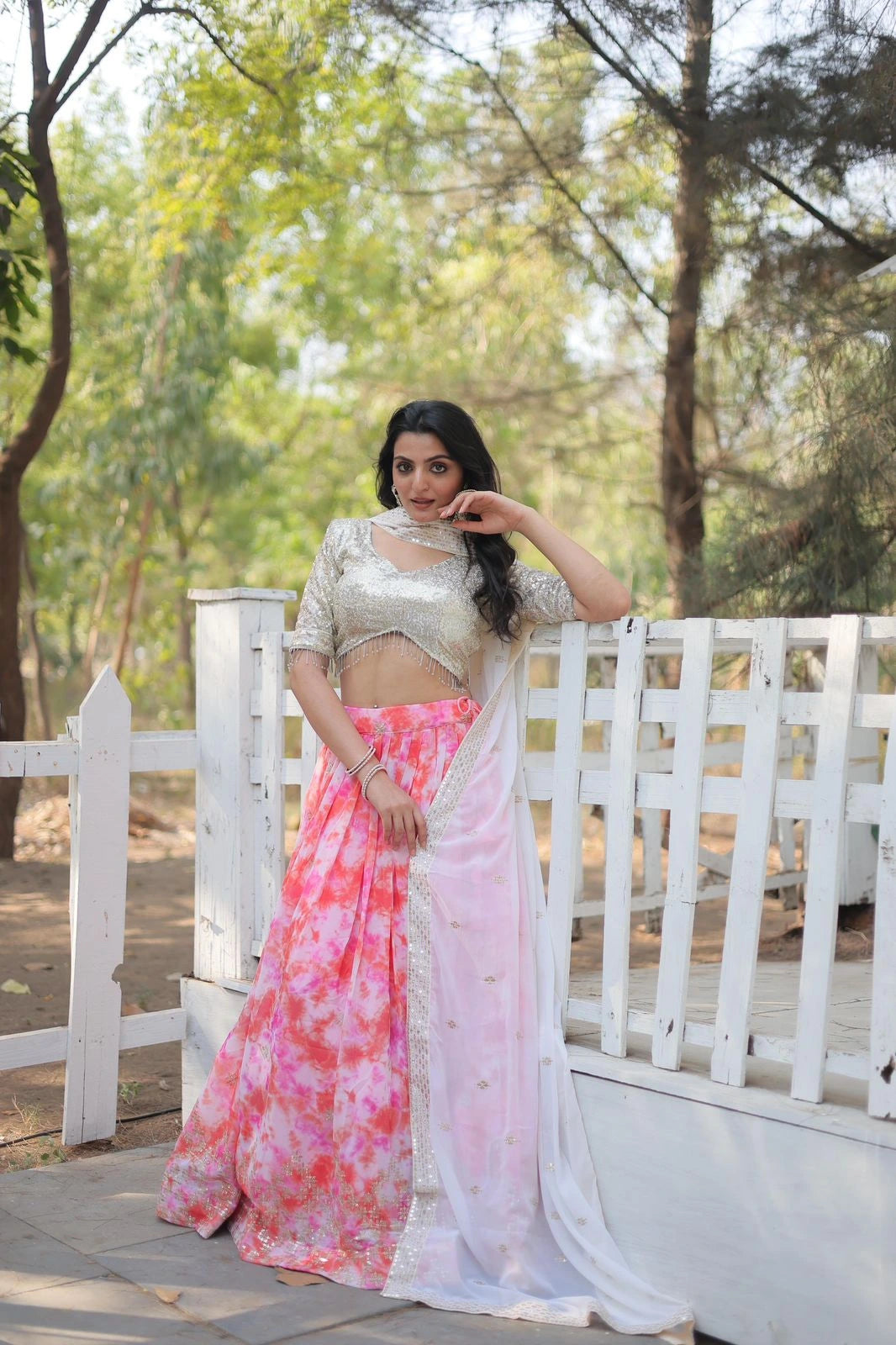 Woman in a pink and white saree standing on a wooden bridge with greenery in the background
