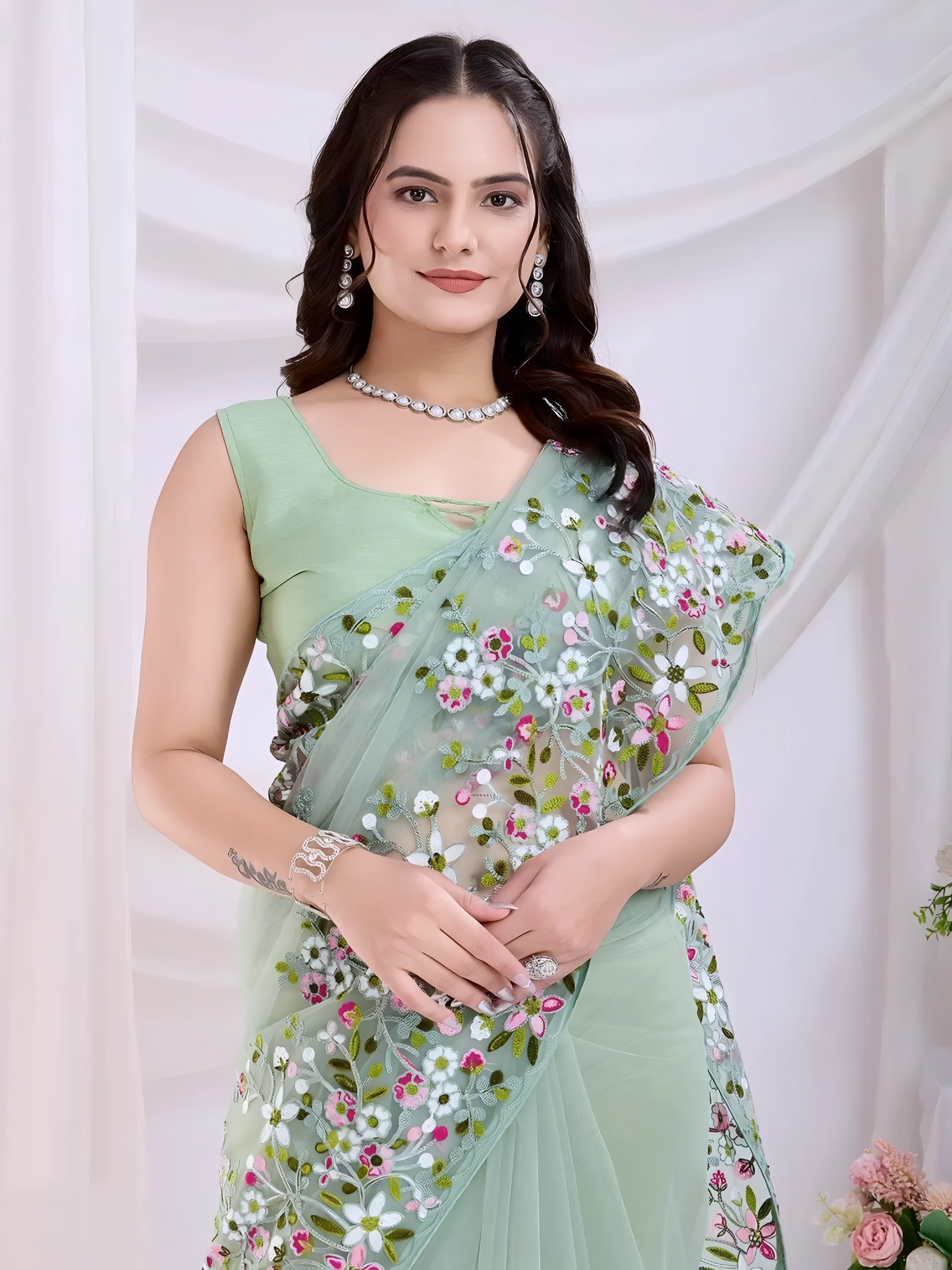 Woman wearing a mint green net saree with intricate multicA woman models an ethereal Light Mint Green Soft Net Saree. The sheer fabric is densely covered in delicate all-over floral embroidery featuring white, pink, and green threadwork. She wears a matching sleeveless light green blouse and accessorizes with delicate silver/diamond jewelry, posing indoors against sheolor floral embroidery and a matching sleeveless blouse, posing in a modern studio setting with white draped curtains and floral arrangements.