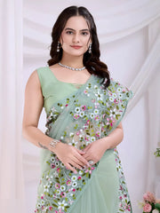 Woman wearing a mint green net saree with intricate multicA woman models an ethereal Light Mint Green Soft Net Saree. The sheer fabric is densely covered in delicate all-over floral embroidery featuring white, pink, and green threadwork. She wears a matching sleeveless light green blouse and accessorizes with delicate silver/diamond jewelry, posing indoors against sheolor floral embroidery and a matching sleeveless blouse, posing in a modern studio setting with white draped curtains and floral arrangements.