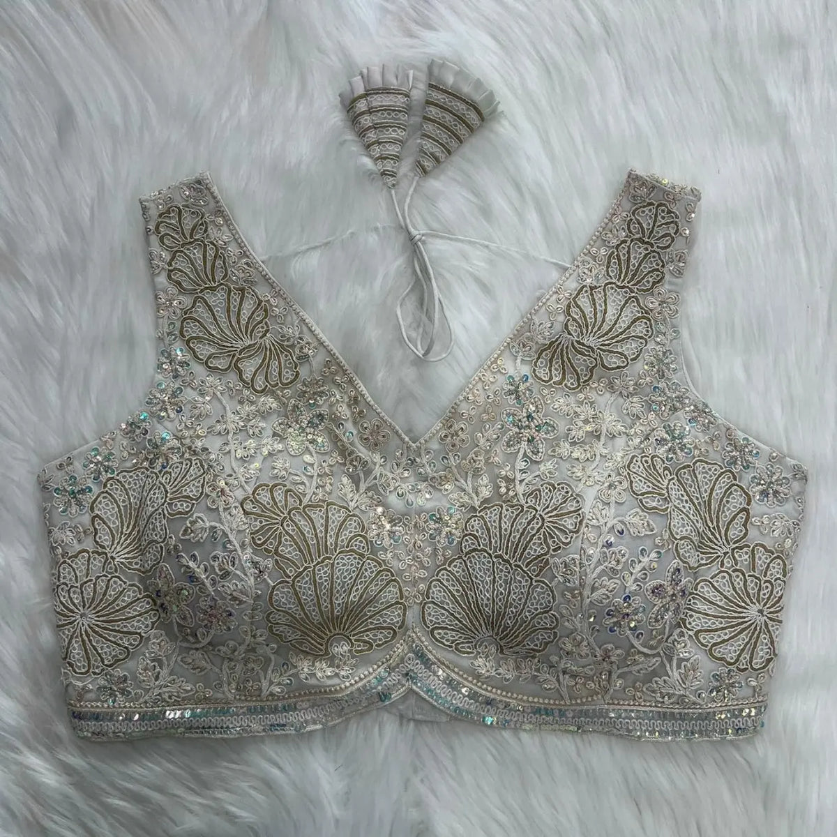Ivory blouse featuring intricate gold sequin and thread embroidery with floral and shell motifs, shimmering bead embellishments, a deep V-neck, structured cups, and decorative tasseled tie-back detail, displayed flat on a white fur background.