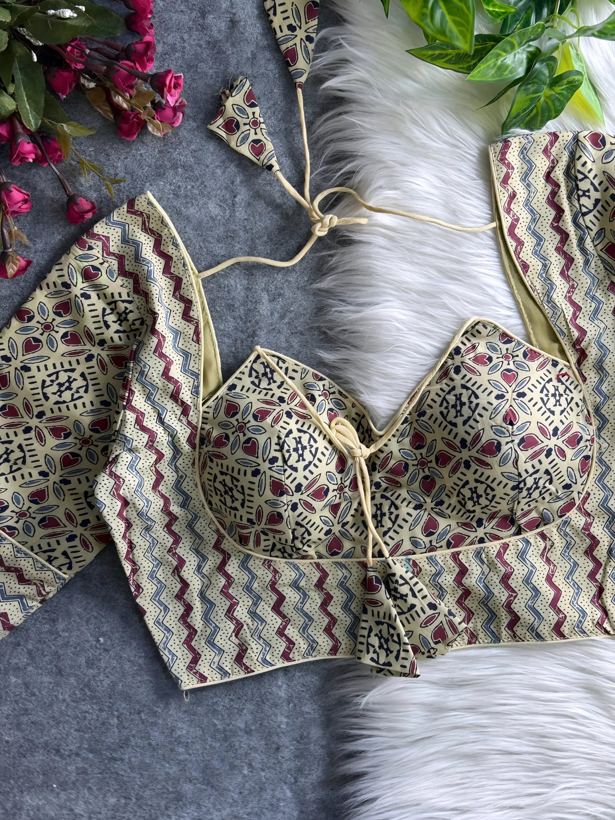 Beige padded blouse with maroon and navy geometric and floral patterns, displayed on a white fur surface with artificial flowers and green leaves in the background.Beige padded blouse with maroon and navy geometric and floral patterns, displayed on a white fur surface with artificial flowers and green leaves in the background.