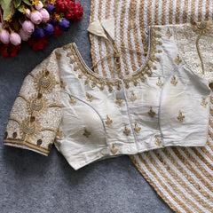 Traditional ivory silk blouse featuring intricate gold embroidery and beadwork with floral and paisley motifs on the sleeves and neckline, displayed flat on a textured surface with a bunch of artificial flowers and a woven mat in the background.